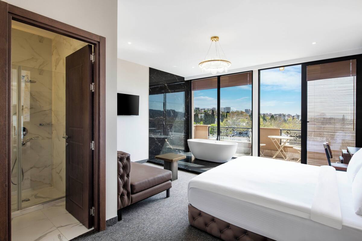 Une chambre moderne dotée d’un grand lit, d’un coin salon, d’une baignoire et d’un balcon offrant une vue sur la ville.