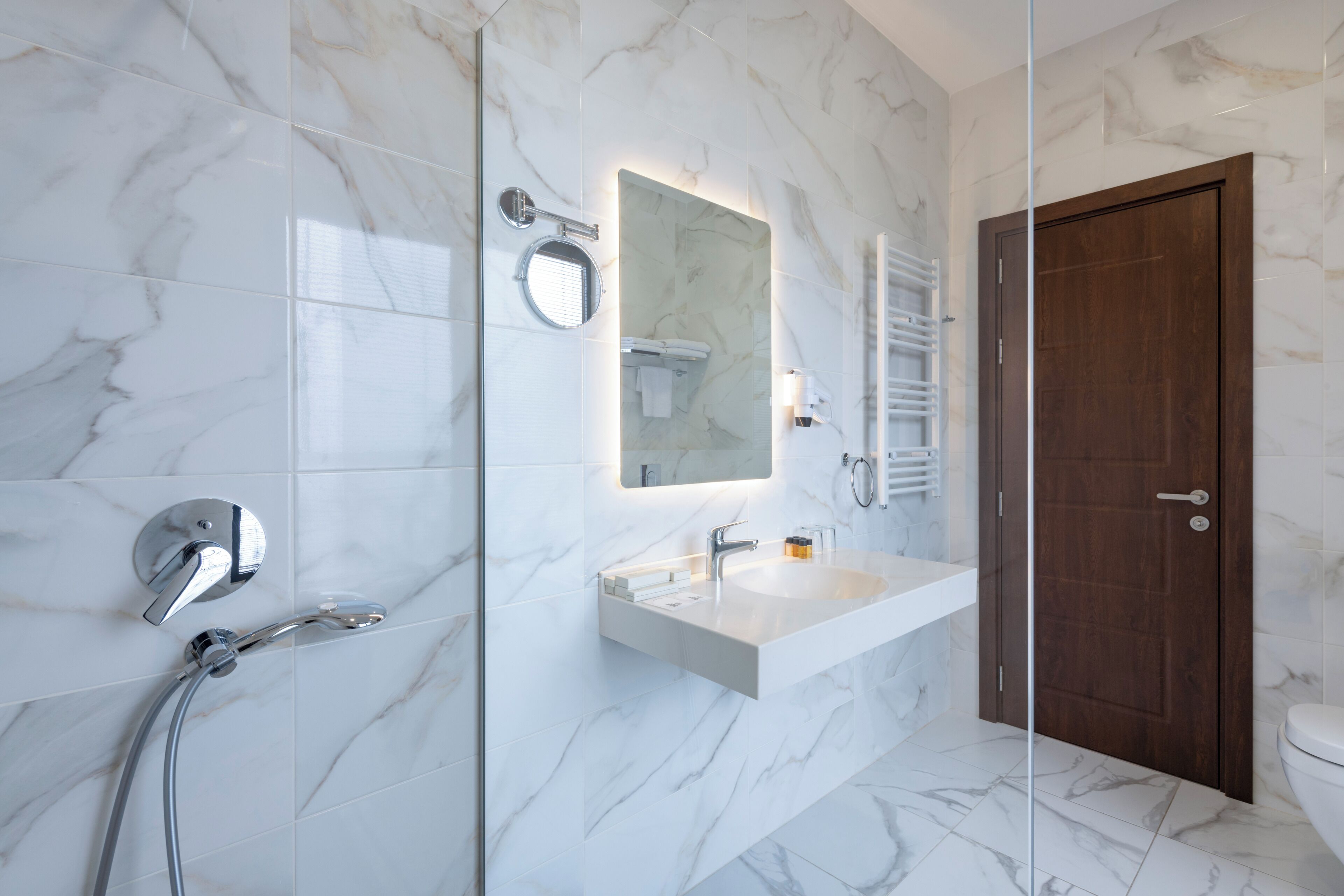 A modern bathroom with a glass shower enclosure, a marble-tiled wall, a white sink, and a wooden door.