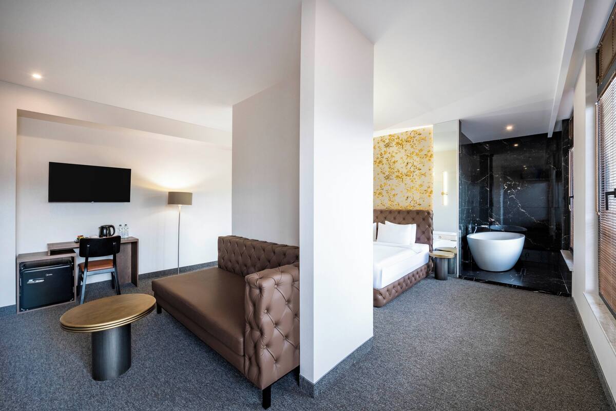 Une chambre d’hôtel équipée d’un canapé, d’un bureau, d’un téléviseur à écran plat, d’un lit, d’une grande baignoire et d’un téléviseur fixé au mur.