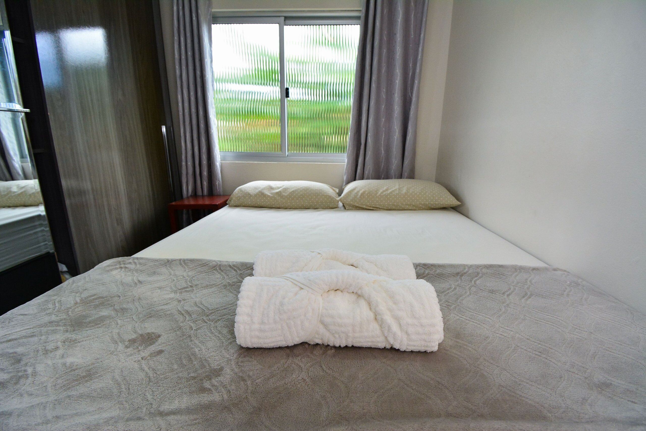 Cama bem arrumada com lençóis brancos e duas almofadas, janela com cortinas e vista para uma paisagem verde.