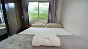 A neatly made bed with white linens and two pillows, a window with curtains, and a view of a green landscape. - Casa Aconchegante na Gamboa - GAR003 (Garopaba)