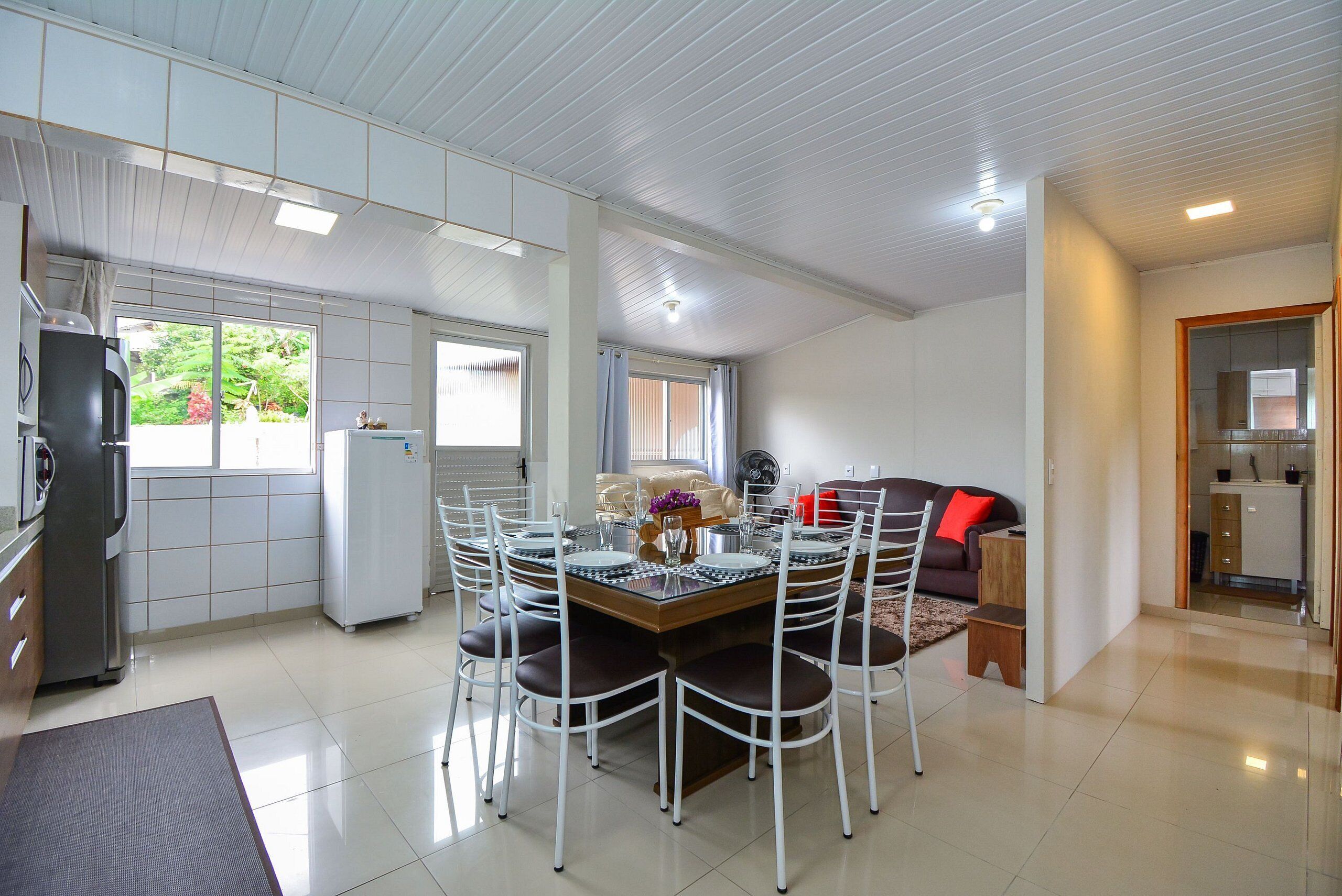 A modern kitchen with a dining area, white tiled floors, and a view of greenery outside.
