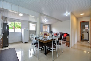 A modern kitchen with a dining area, white tiled floors, and a view of greenery outside.