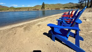 Een rij blauwe Adirondack-stoelen op een zandstrand aan een rustige rivier, met bergen op de achtergrond.