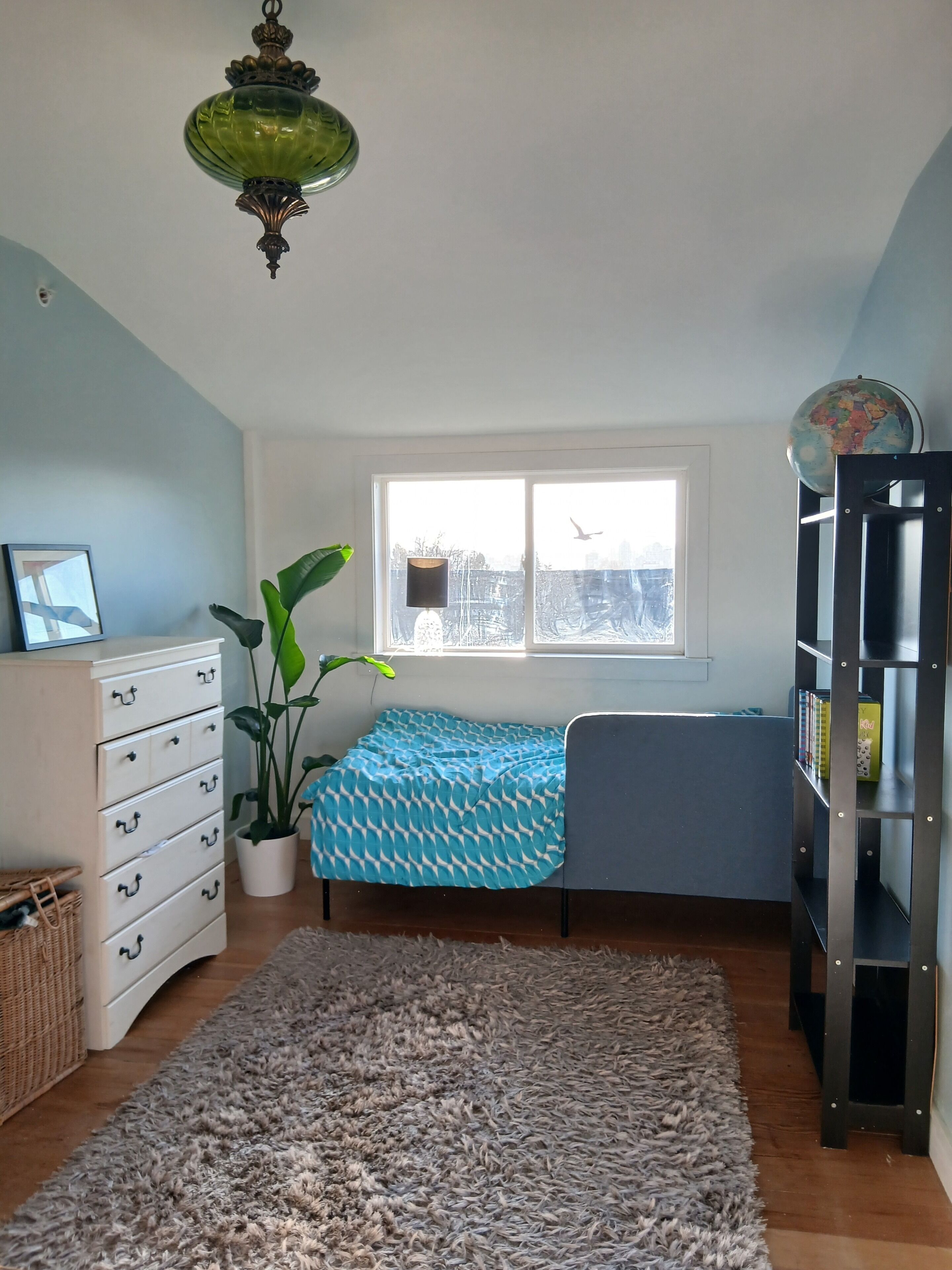 A room with a bed, dresser, potted plant, bookshelf, and a window with a view of trees.