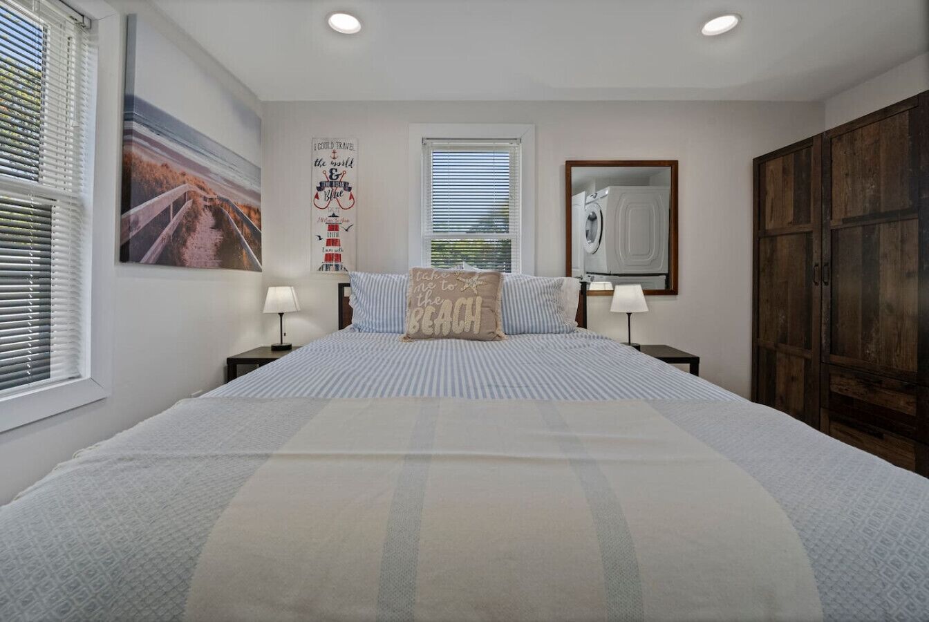 Une chambre à coucher avec un grand lit, deux tables de chevet, une fenêtre avec des stores, un miroir fixé au mur et une armoire en bois.