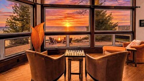 Une pièce dotée d’une grande fenêtre donnant sur un coucher de soleil, de deux fauteuils en cuir, d’une table de baby-foot et d’une petite table avec des chaises.