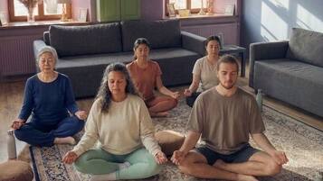 Un grupo de personas meditando en una habitación con un tapete estampado, un armario verde y un sofá gris.