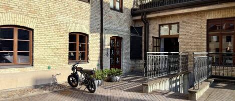 A brick building with a motorcycle parked in front, a balcony with a wrought iron railing, and a paved courtyard.