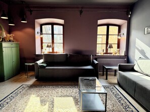 Ett modernt vardagsrum med en soffa, ett soffbord och ett element. Det finns två fönster med gardiner, ett grönt skåp och en mönstrad matta.