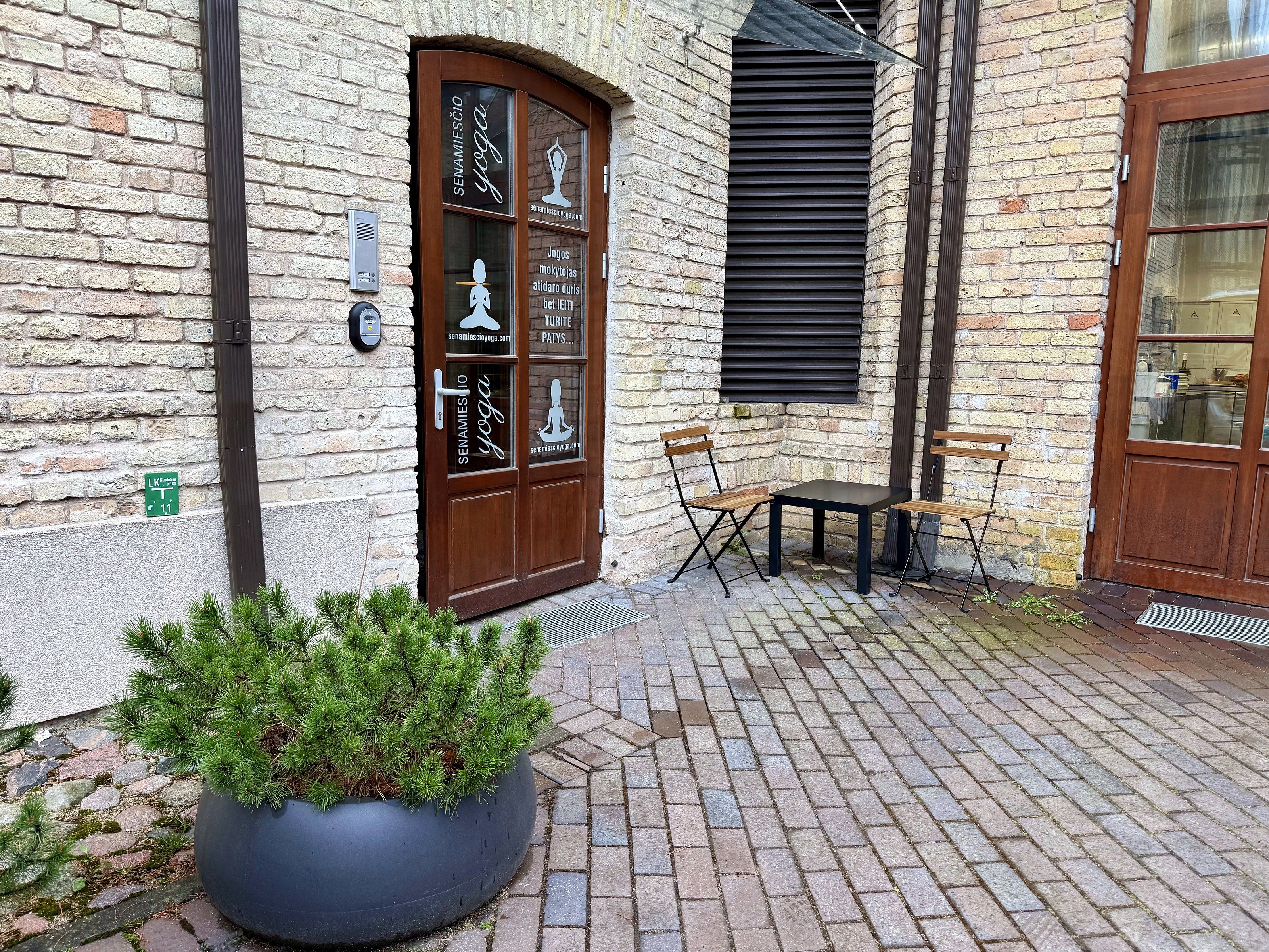 A brick building with a wooden door labeled 'SENAMIESIO yoga,' a small outdoor seating area, and a potted plant.
