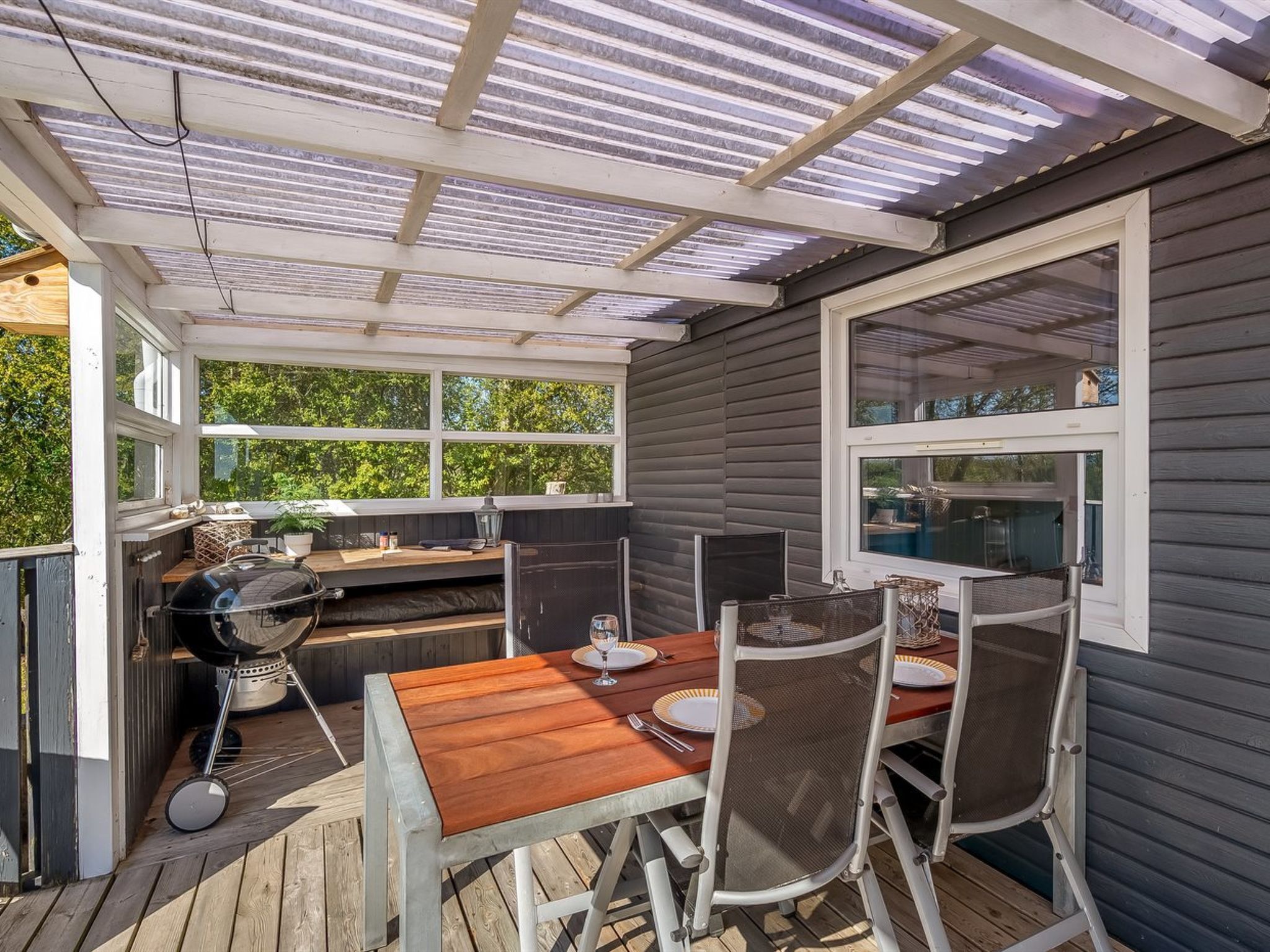 Un patio exterior con mesa y sillas de madera, una parrilla y una ventana con vistas a la vegetación.