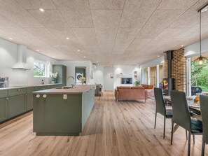 Eine moderne Küche mit einer zentralen Kochinsel, ein Essbereich mit Stühlen und ein Wohnbereich mit Sofa und Fernseher.