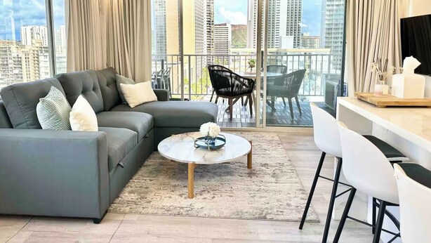 Uma sala de estar moderna com um sofá modular cinza, uma mesa de centro redonda e uma varanda com mobiliário para área externa.