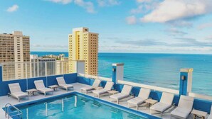 Piscina no terraço com espreguiçadeiras, desenho de borboleta na água e vista para o oceano e edifícios.