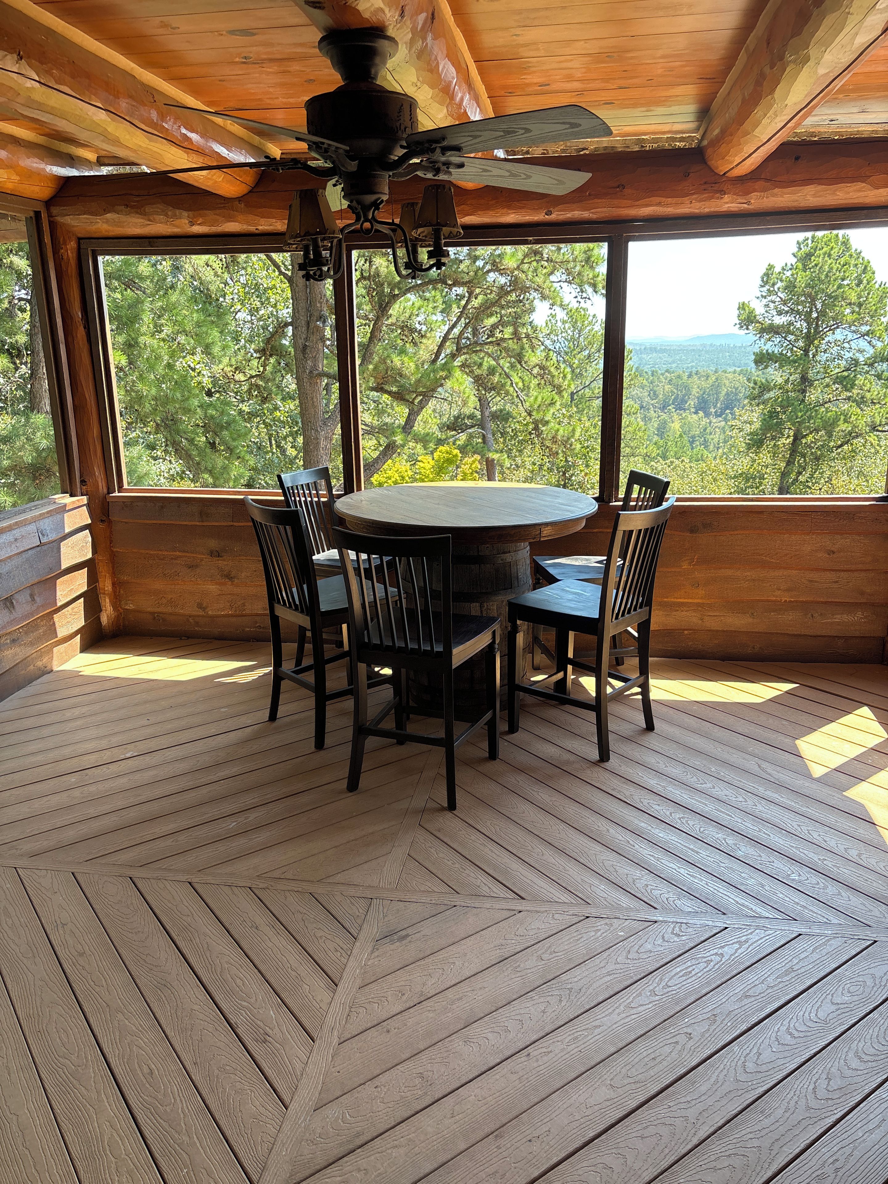 A wooden deck with a round table and chairs, large windows with a forest view, and a ceiling fan.