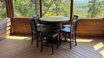 A wooden deck with a round table and chairs, large windows with a forest view, and a ceiling fan.