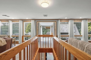 A spacious interior with a wooden staircase leading to a balcony, large windows, and a view of greenery outside.