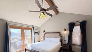 Un dormitorio con cama, ventilador de techo, mesita de noche, ventana con cortinas y vistas al exterior.