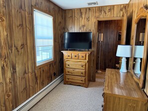 Ein Zimmer mit Holzvertäfelung, einem Flachbildfernseher auf einer Holzkommode, einem Fenster mit Jalousien, einem Spiegel und einer Lampe.