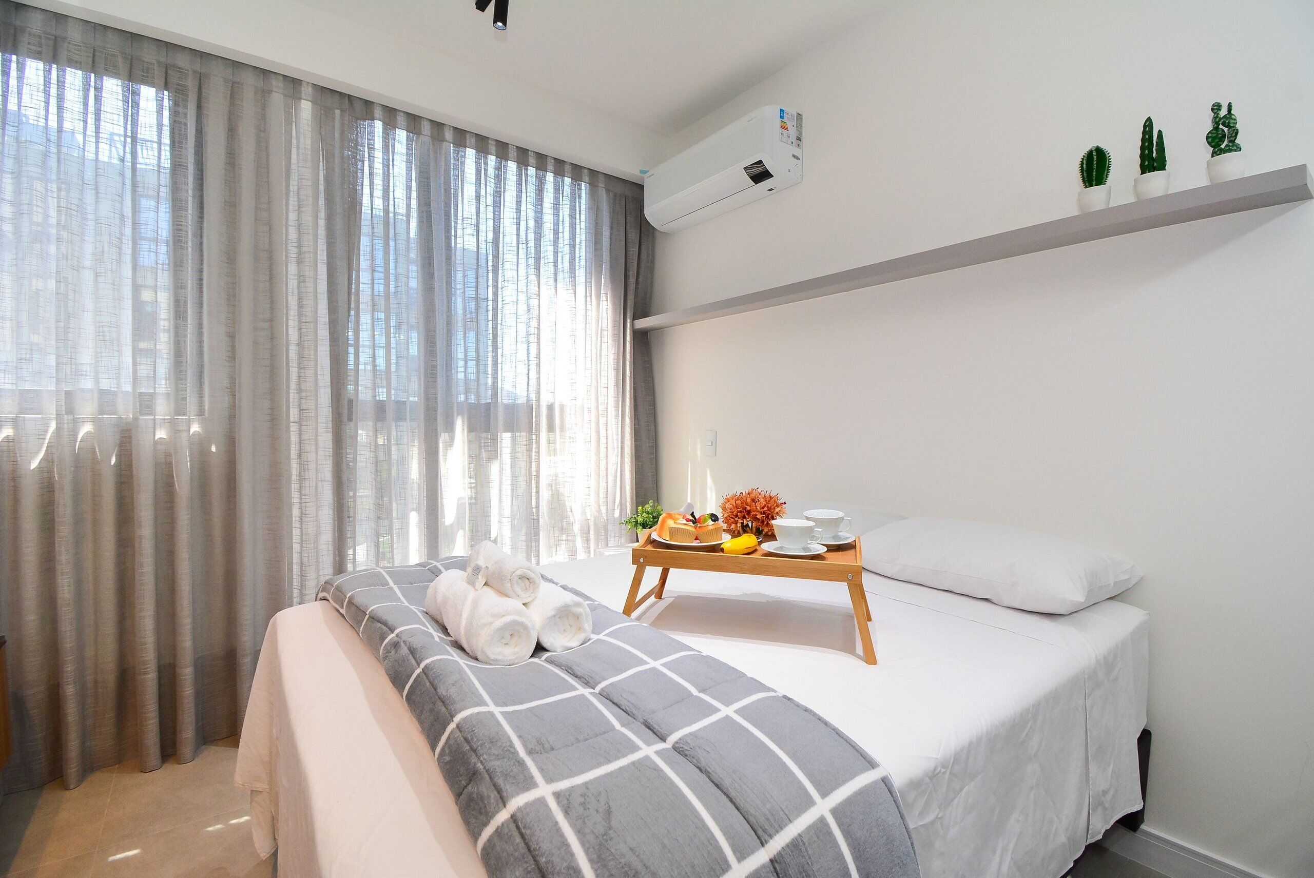 A hotel room with a bed, a bedside table with a tray of fruit and a cup, a window with curtains, and an air conditioning unit.