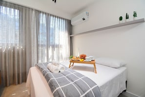 Une chambre d’hôtel avec un lit, une table de chevet sur laquelle se trouve une assiette de fruits et une tasse, une fenêtre avec des rideaux et un appareil de climatisation.
