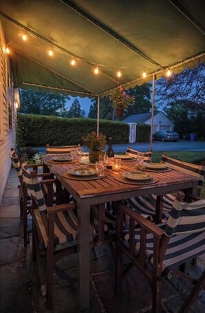 Een terras buiten met een houten eettafel, omringd door gestreepte stoelen, onder een luifel met lichtslingers.
