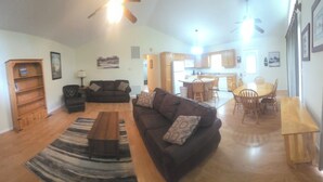 Sala de estar con sofá, mesa de centro, televisor, estantería y área de cocina al fondo.