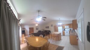 Amplia sala de estar con mesa de comedor, sillas, sofá, ventilador de techo y cocina equipada con armarios y electrodomésticos.