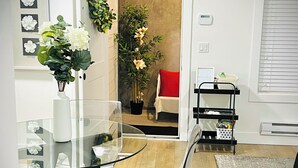 A modern living room with a glass dining table, clear chairs, a red sofa, a black chair, a small black shelf, and a potted plant. - Bright 2BR Suite Near White Rock & Vancouver (Surrey)