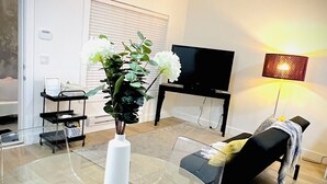 A modern living room with a glass table, a white vase with flowers, a black sofa, a TV, and a floor lamp. - Bright 2BR Suite Near White Rock & Vancouver (Surrey)