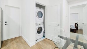 A laundry room with a front-loading washer and dryer, a glass table, and a bed with a patterned bedspread in the adjacent room. - Bright 2BR Suite Near White Rock & Vancouver (Surrey)