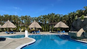 Ein Swimmingpool mit Liegestühlen und Strohdächern, umgeben von Palmen.
