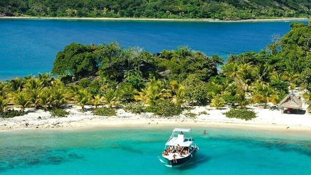 Ein Boot auf einem türkisblauen Meer mit Strand und bewaldetem Hügel im Hintergrund.