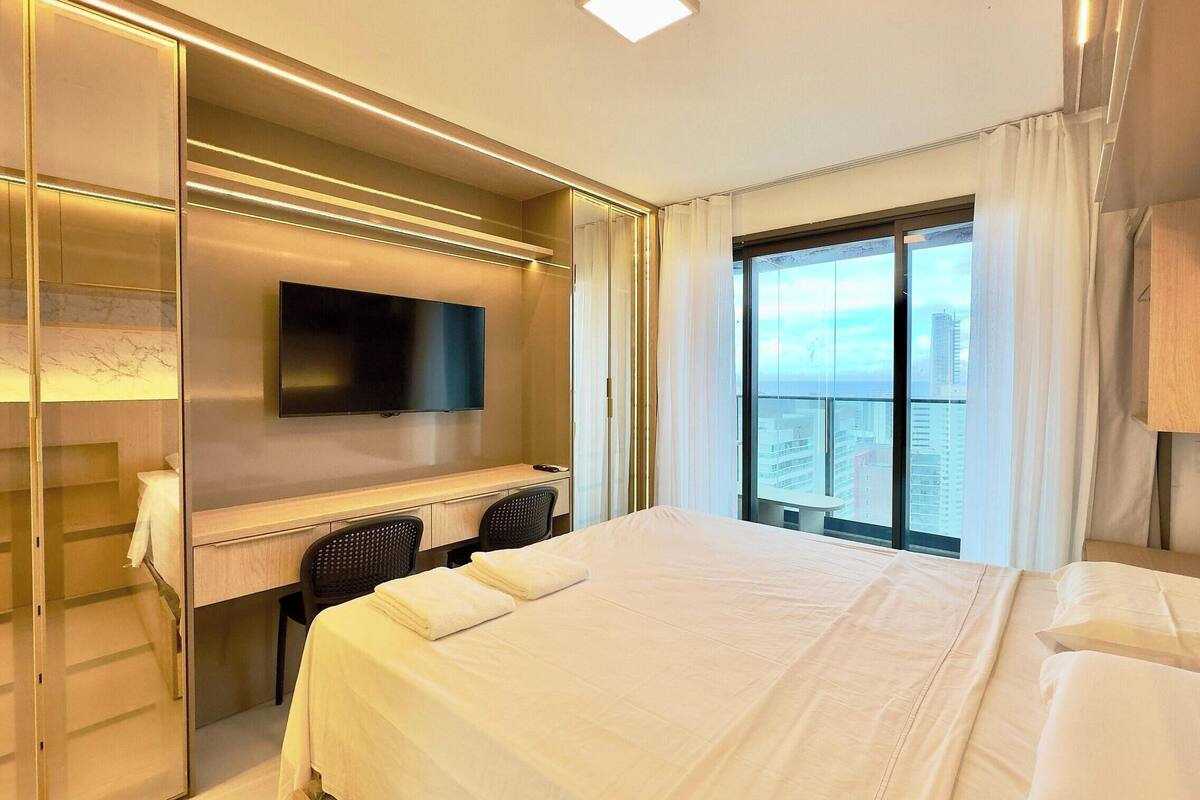 A hotel room with a bed, a desk with a chair, a TV mounted on the wall, and a large window with a city view.