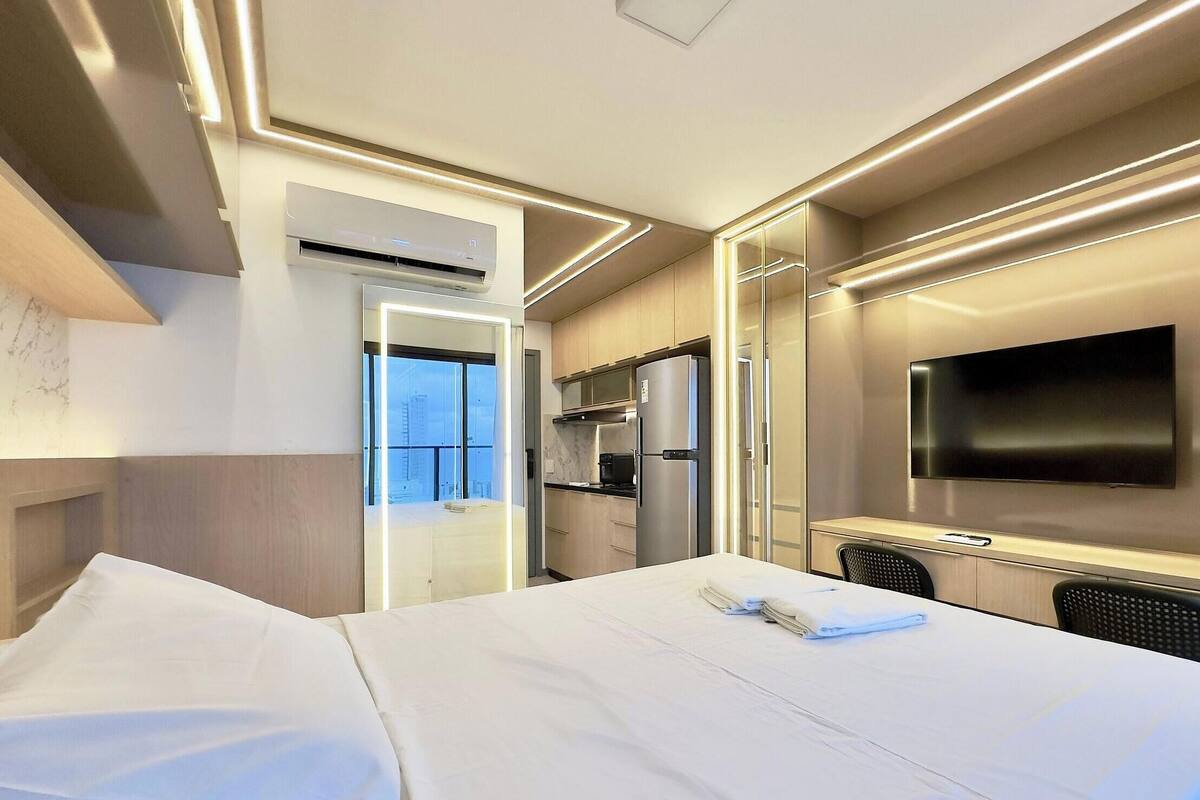A hotel room with a bed, a TV, a kitchenette, and a view of the city.