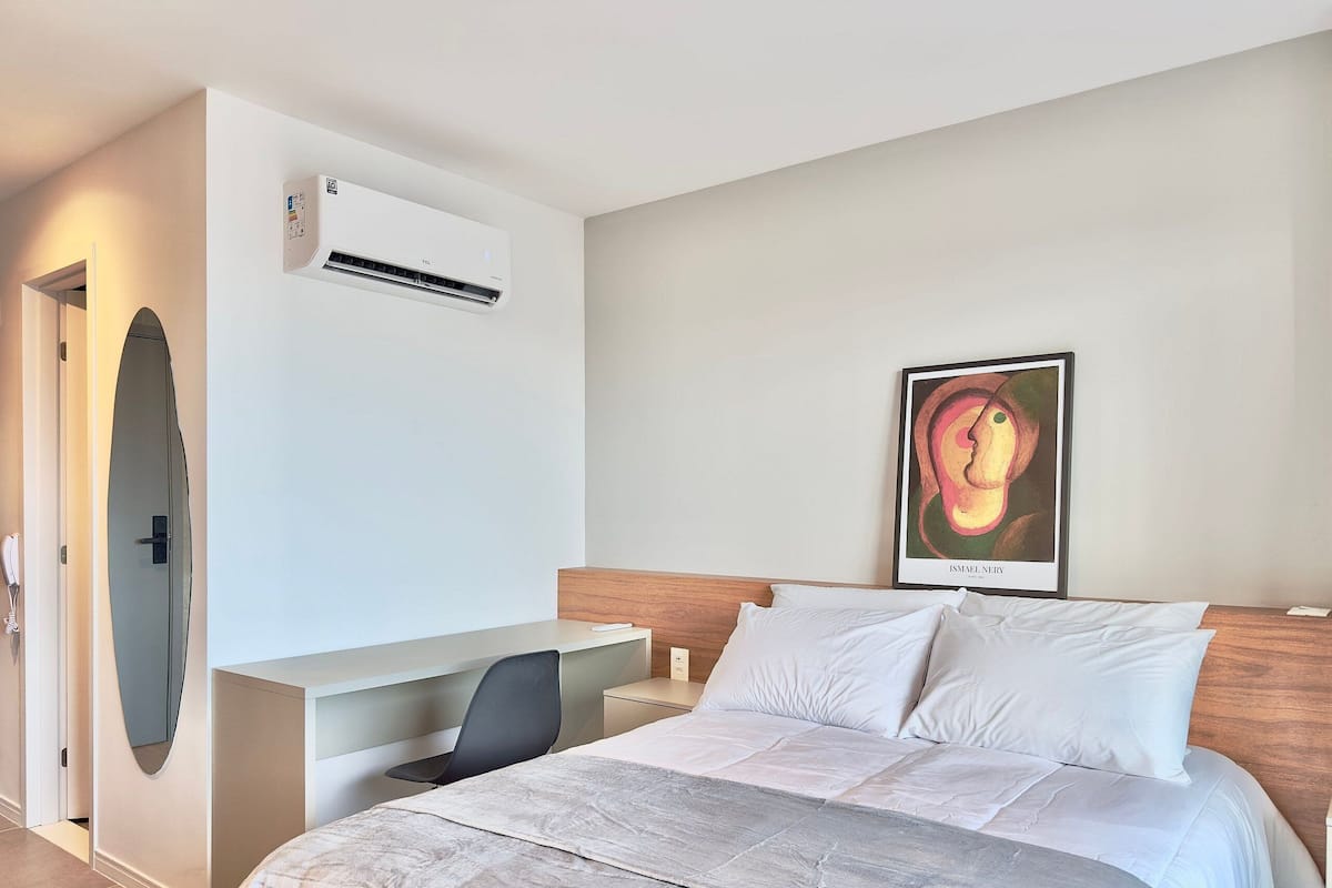 A hotel room with a bed, a desk, a chair, an air conditioning unit, and a framed artwork on the wall.