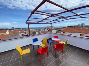 Eine Dachterrasse mit rotem Metallgeländer, einem Tisch mit Stühlen und Blick auf die Stadt.