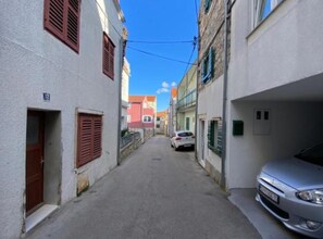 Eine schmale Straße mit Gebäuden an beiden Seiten, ein Auto parkt rechts, und ein klarer, blauer Himmel.