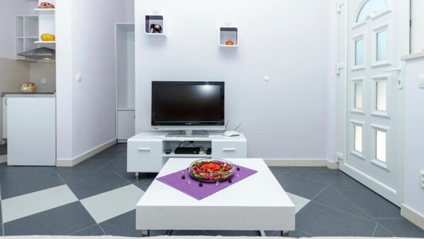 Una sala de estar moderna con un televisor de pantalla plana, una mesa de comedor con un mantel individual morado, y una zona de cocina con armarios blancos y un refrigerador.