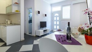 Una cocina moderna con armarios blancos, una zona de comedor con mesa y sillas blancas, un televisor y una orquídea en maceta.