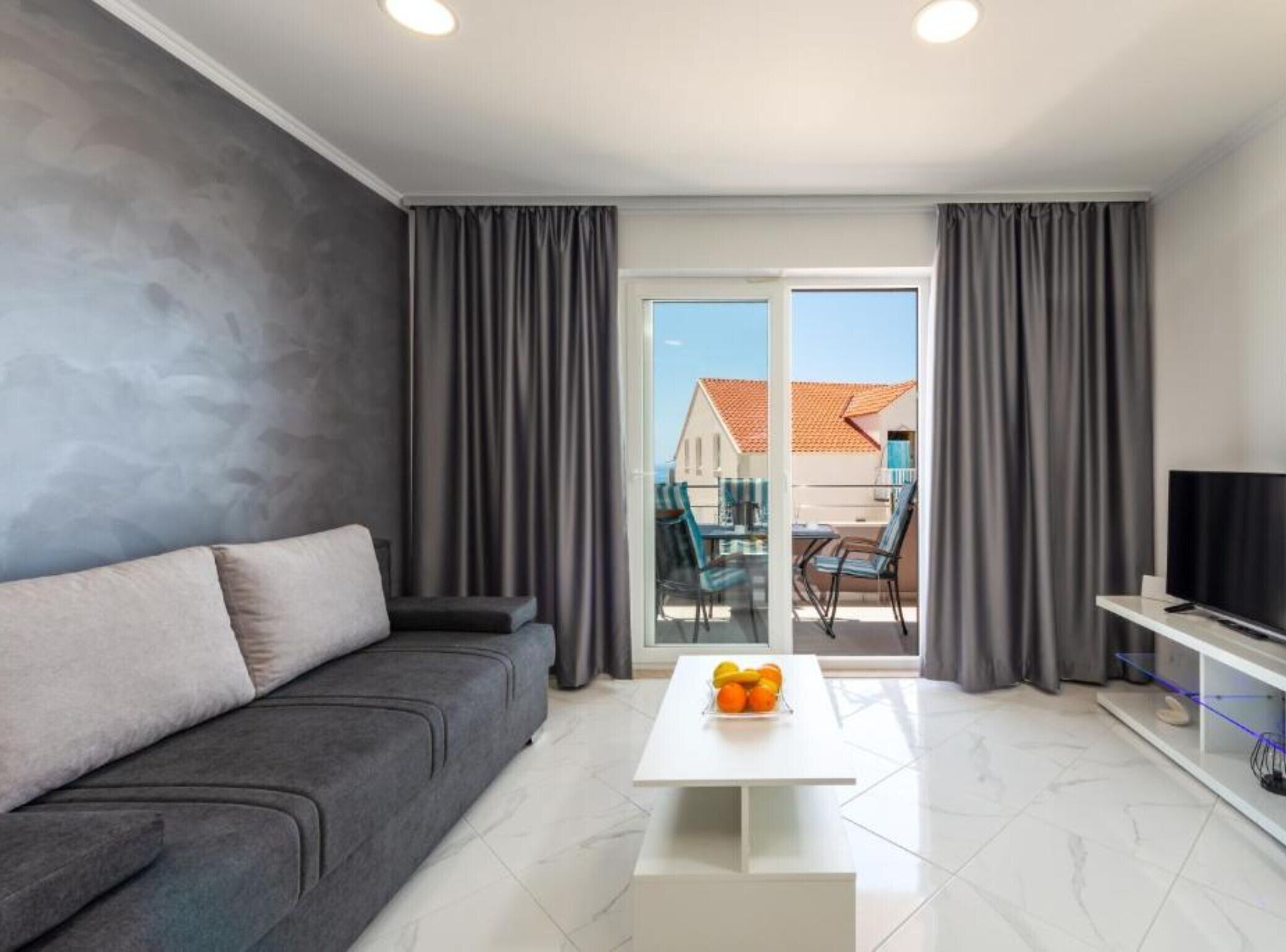 Ein modernes Wohnzimmer mit einem grauen Sofa, einem weißen Couchtisch mit Früchten und einem Fernseher auf einem Schrank.