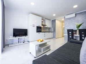 Un soggiorno moderno con una TV a schermo piatto, un tavolino da caffè bianco con frutta, un divano grigio e un angolo cottura con pensili bianchi e un forno nero.