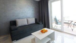 Een moderne woonkamer met een donkergrijze bank, een witte salontafel met fruit, en een grote schuifdeur naar een buitenruimte met stoelen.