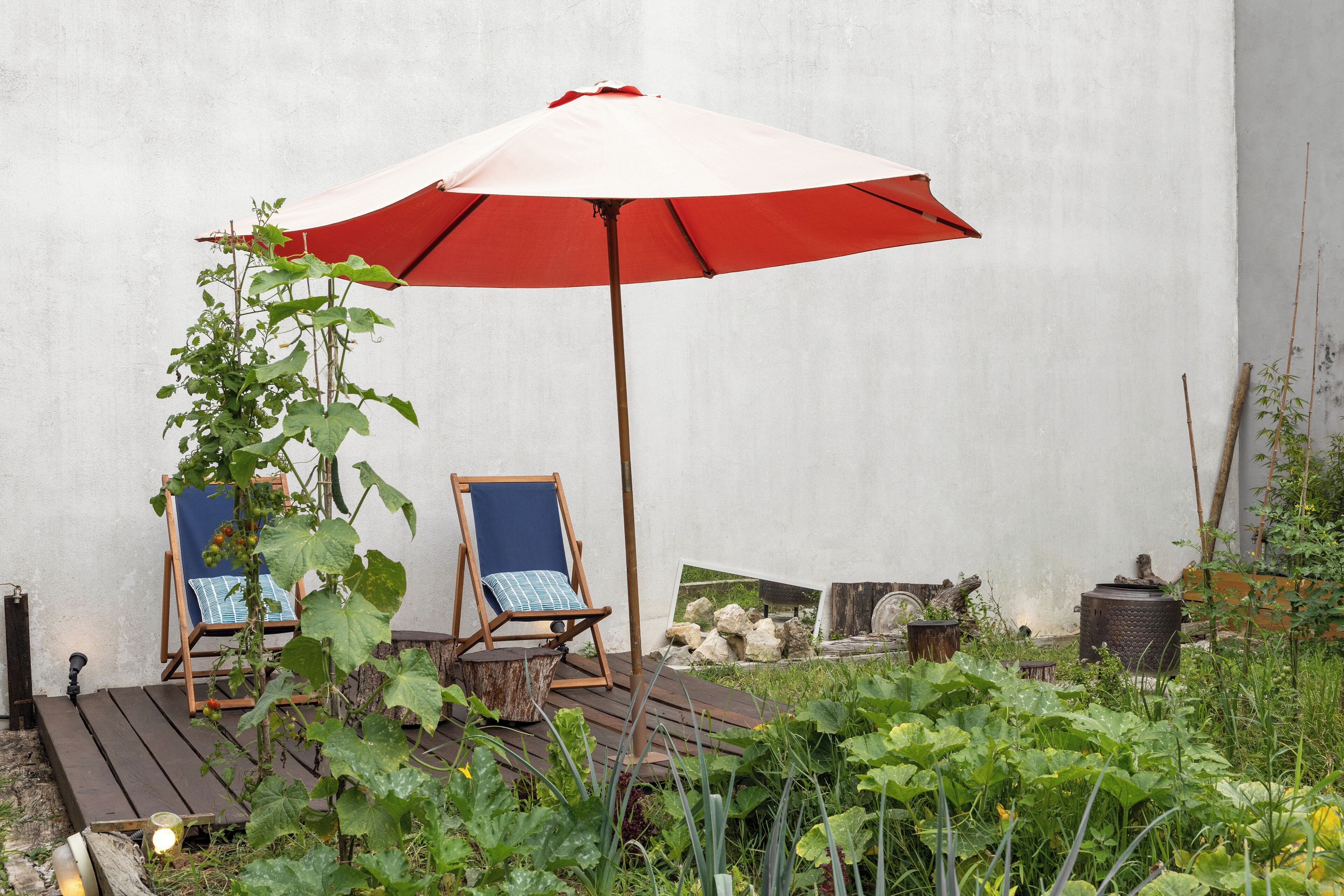 A wooden deck with two blue chairs under a red umbrella, surrounded by greenery and a white wall.