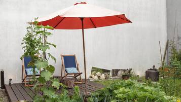 A wooden deck with two blue chairs under a red umbrella, surrounded by greenery and a white wall.
