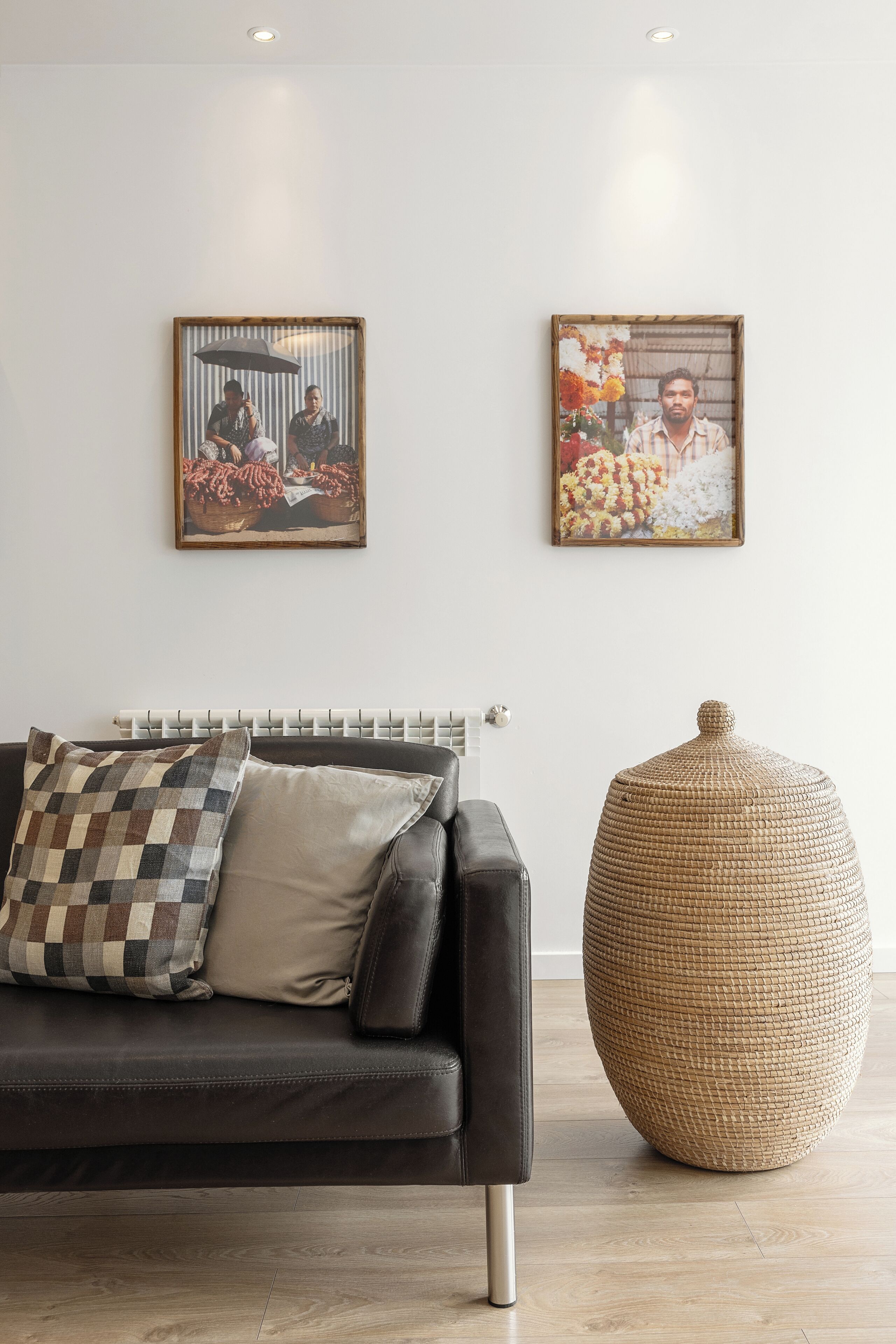 Una habitación con un sofá de cuero negro, dos cuadros en la pared, un radiador y un otoman tejido.