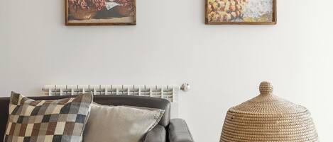 Una habitación con un sofá de cuero negro, dos cuadros en la pared, un radiador y un otoman tejido.
