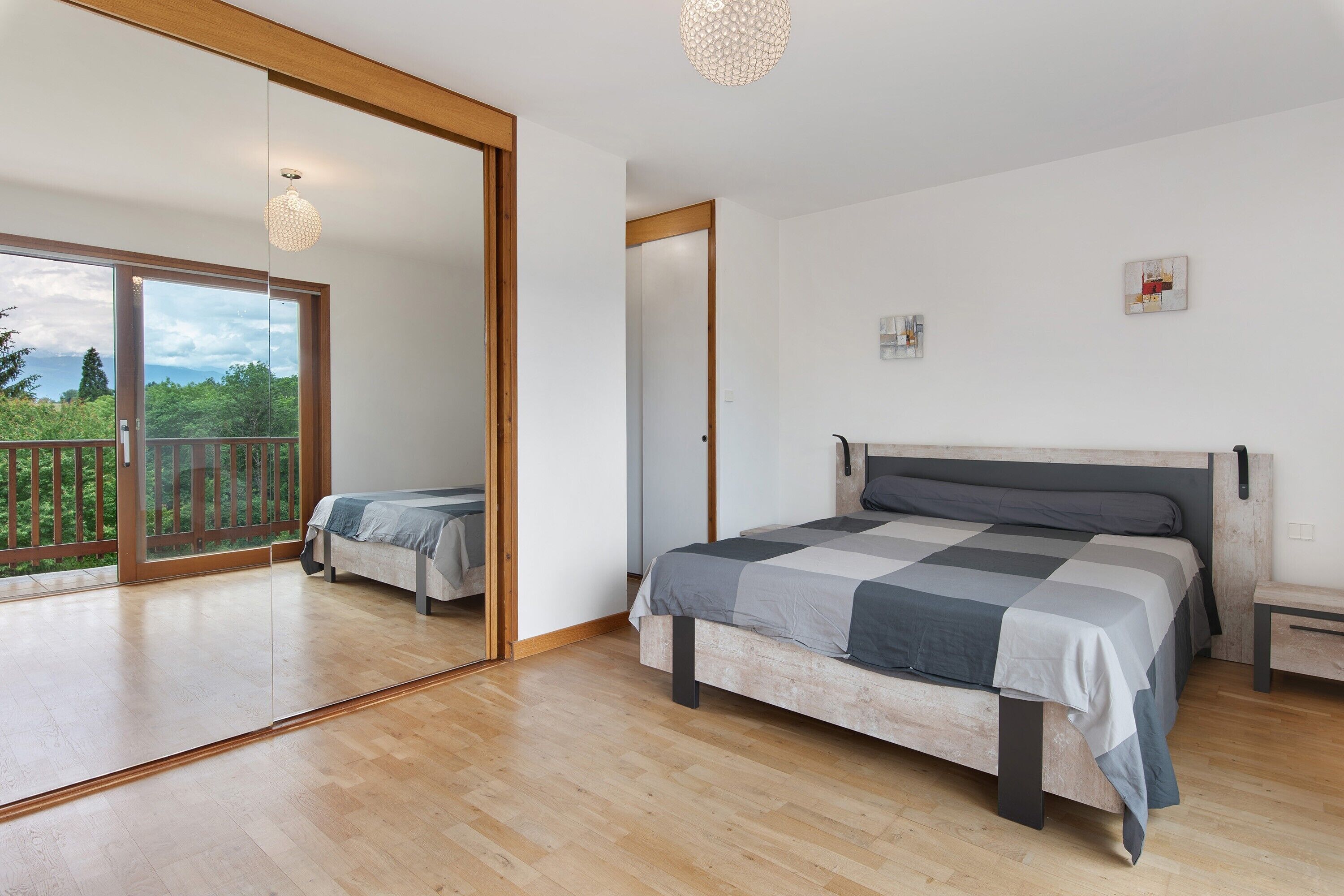 A bedroom with a large bed, a wooden floor, a sliding glass door to a balcony, and a ceiling light fixture.