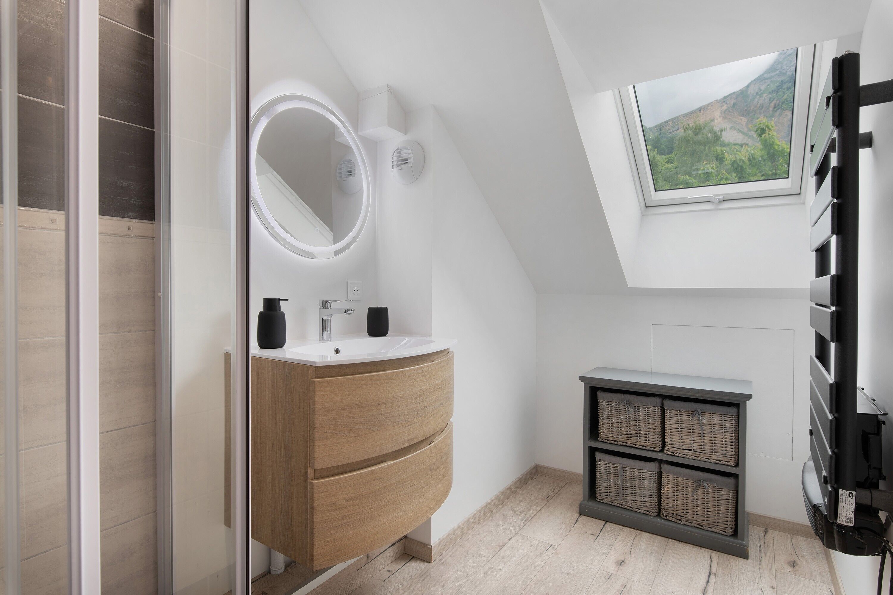 A modern bathroom with a skylight, a round mirror, a sink, and a small cabinet with baskets.